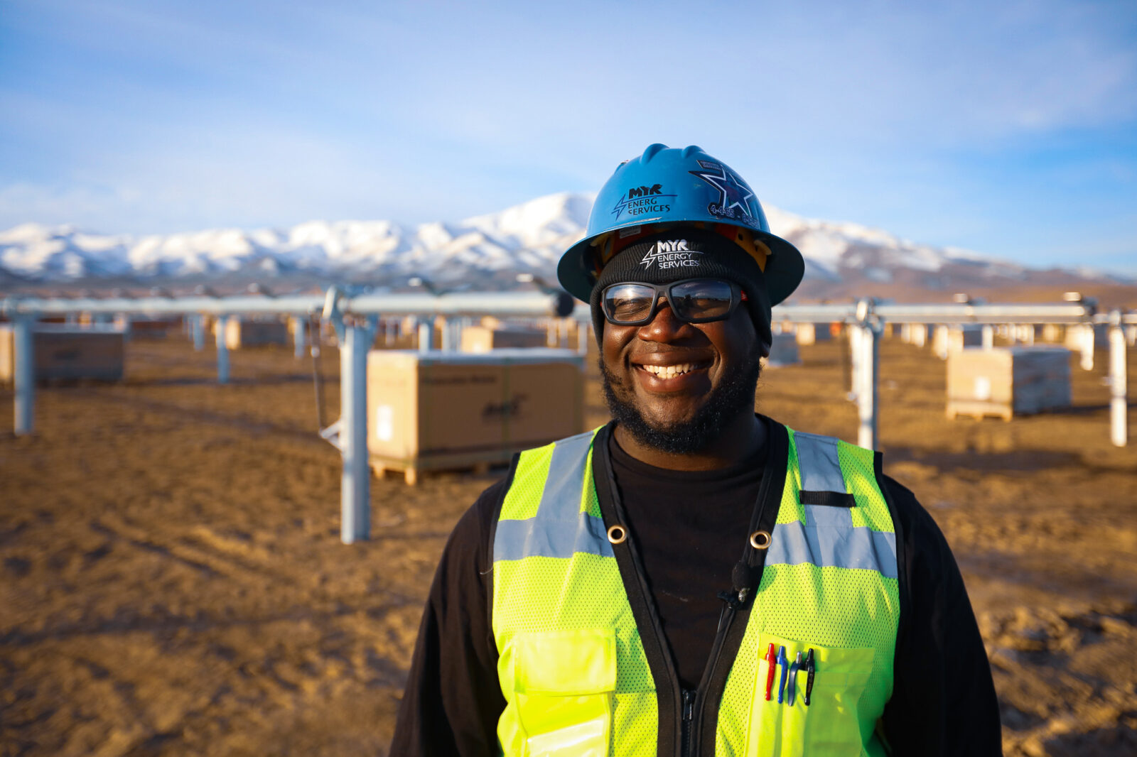 ͵E Energy employee with hard hat and safety glasses smiles for the camera with snowcapped mountains in the distance