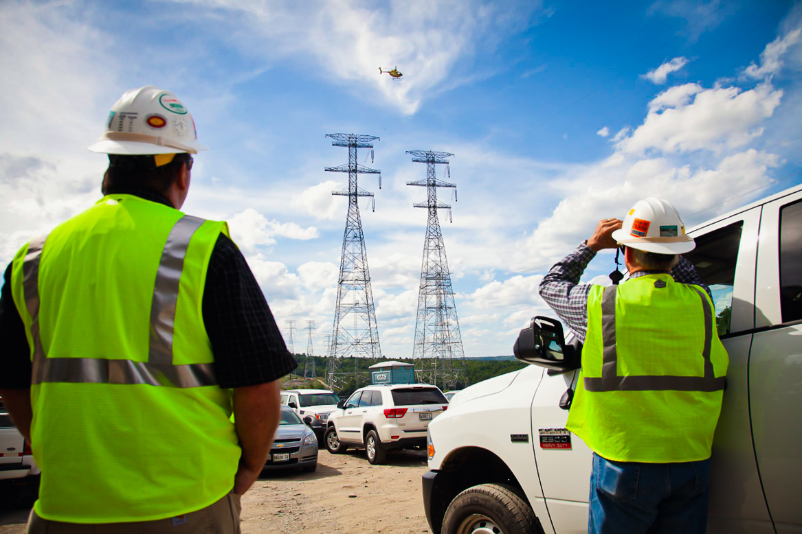 Two ͵ Group employees watch helicopter flying over transmission lines through binoculars